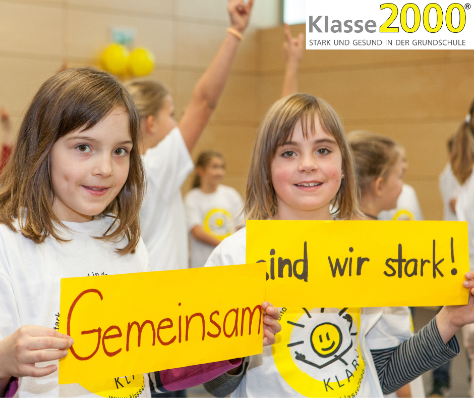 Bild vom Programm Klasse 2000, Es sind 2 Kinder zu sehen, die die Schilder "Gemeinsam" und "sind wir stark!" in die Kamera halten und lächeln.