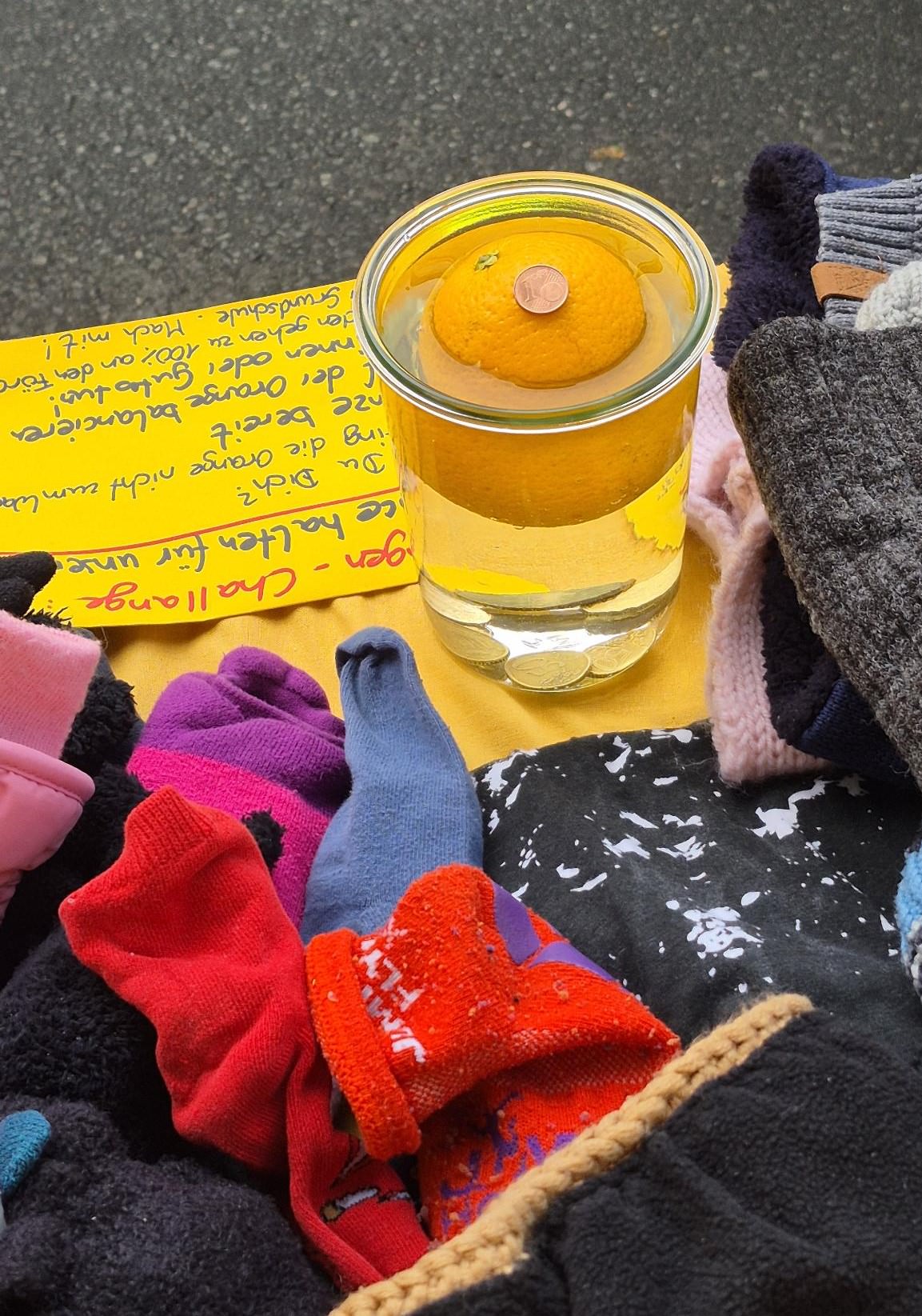 Es ist ein Glas zu sehen, das fast bis zum Rand mit Wasser gefüllt ist. Eine große Orange schwimmt im Wasser und auf der Orange liegt ein 1-Cent-Stück.